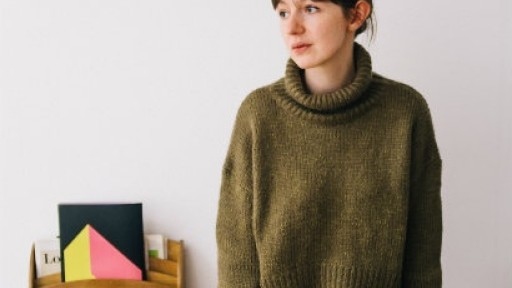 Image of Sally Rooney sitting on the edge of a desk, wearing a green woollen jumper and orange skirt