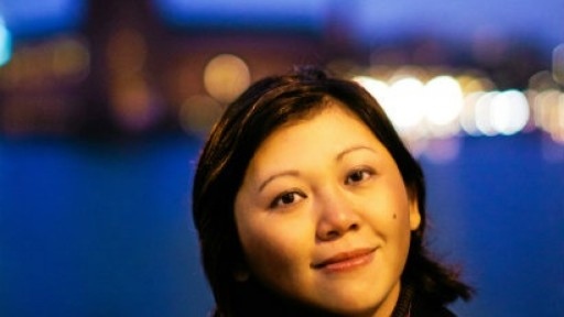 Image of Yiyun Li in a black coat and pink scard, with the hazy lights of a city behind her