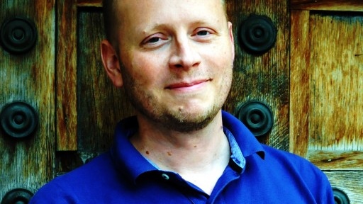 Acclaimed Young Adult author Patrick Ness in a royal blue top, standing infront of an elaborate wooden wall.